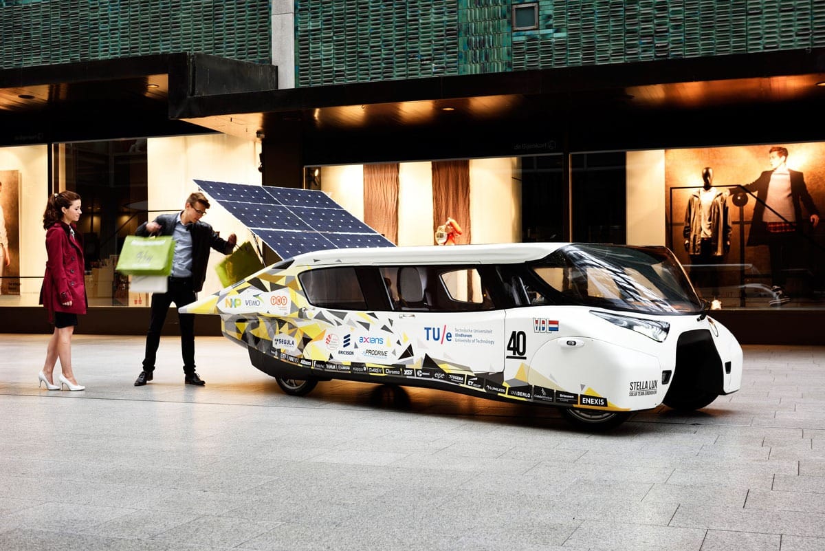 A man loading shopping bags into the back of a mostly white solar car while a woman watches